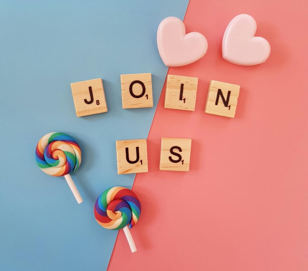 An image of Scrabble tiles which spell out the words Join Us, accompanied by two pink hearts and two lollipops on a blue and salmon pink background.