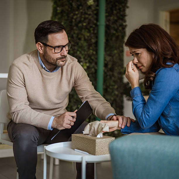 An image of a male psychologist listening intently to a distressed young woman covering her face with a tissue, offering support and guidance in a private counselling session about mental health