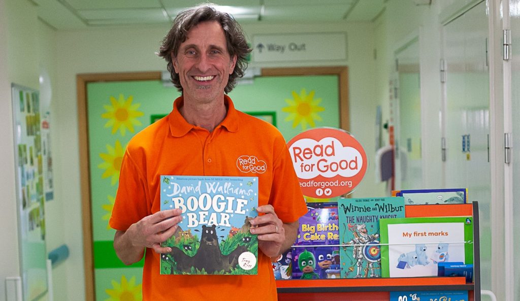 An image of Michael Loader, Read for Good's storyteller, holding a book with a selection of books on a trolley behind him.