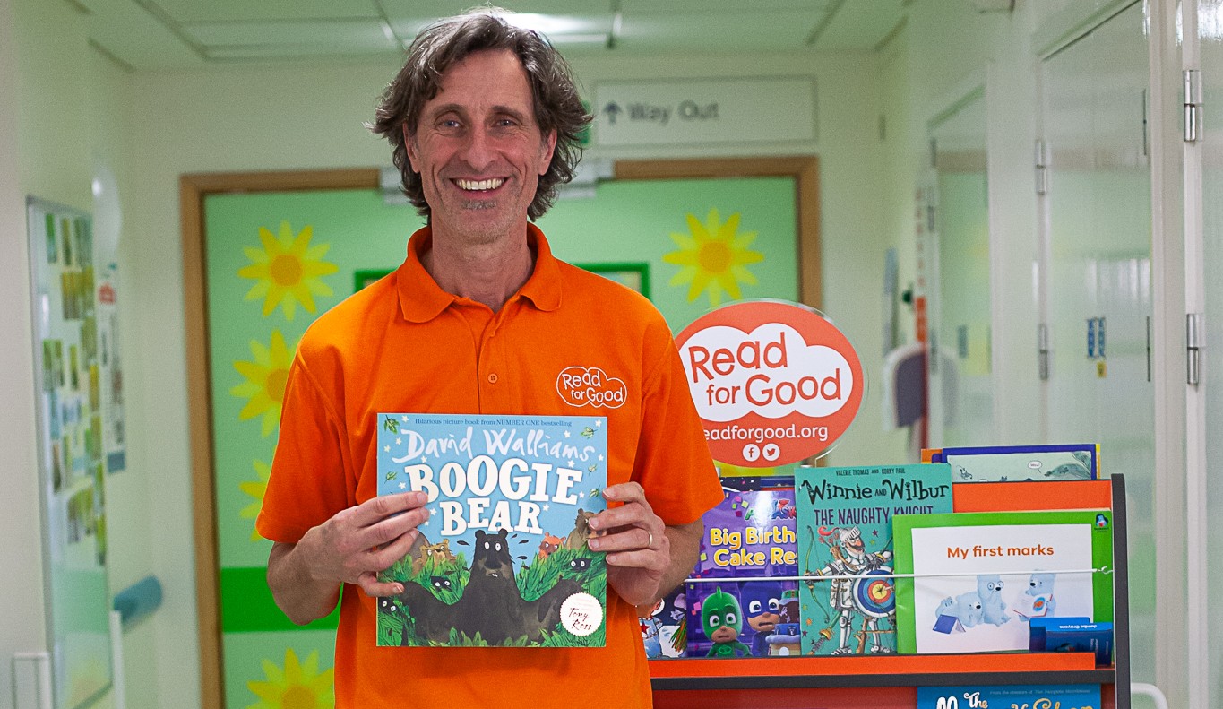 An image of Michael Loader, Read for Good's storyteller, holding a book with a selection of books on a trolley behind him.