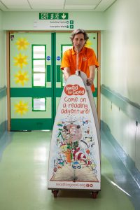 An image of Michael Loader, Read for Good's storyteller pushing a bookcase along a corridor at Royal Cornwall Hospital, Treliske