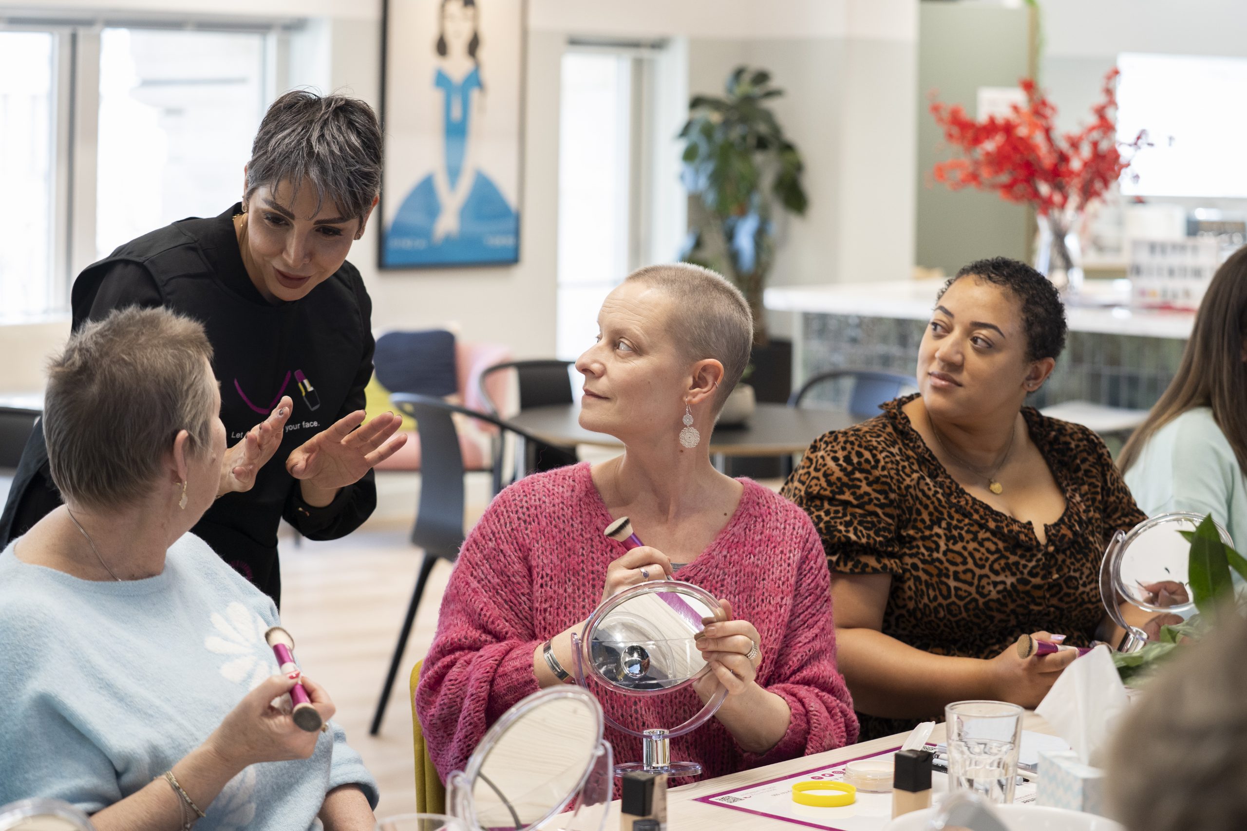 An image showing ladies sat at a table holding mirrors and cosmetics brushes, trying out make up at one of the Look Good Feel Better workshops